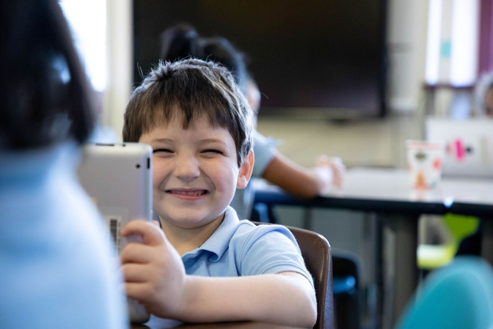 A young student smiles from behind a tablet computer